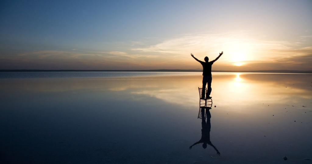Person standing on a chair in the ocean at sunrise with hands raised in prayer